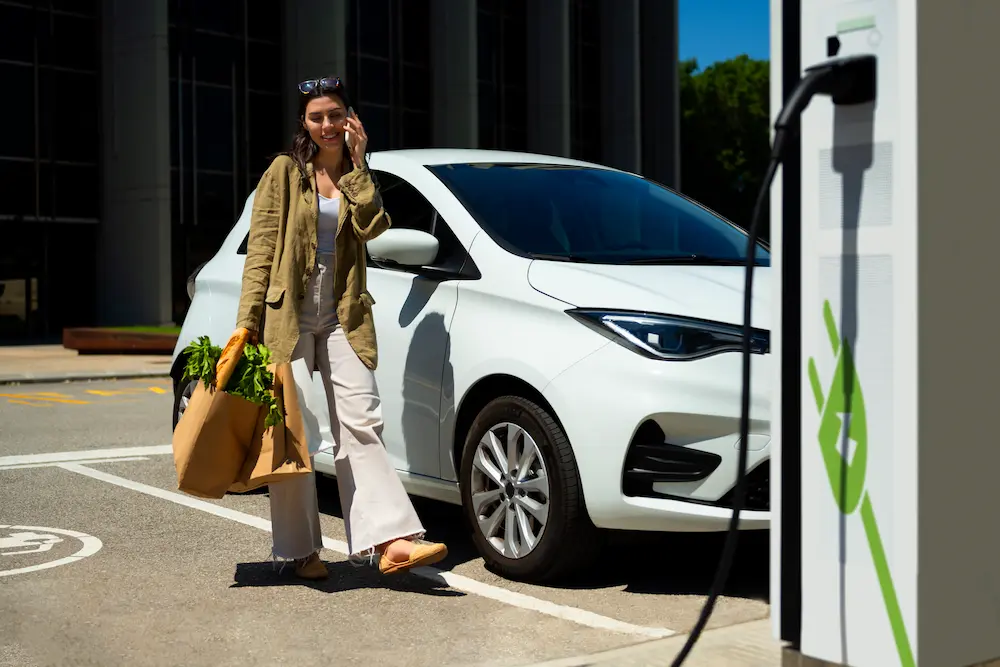 Woman with groceries walking away from an electric car charging at a public EV station.